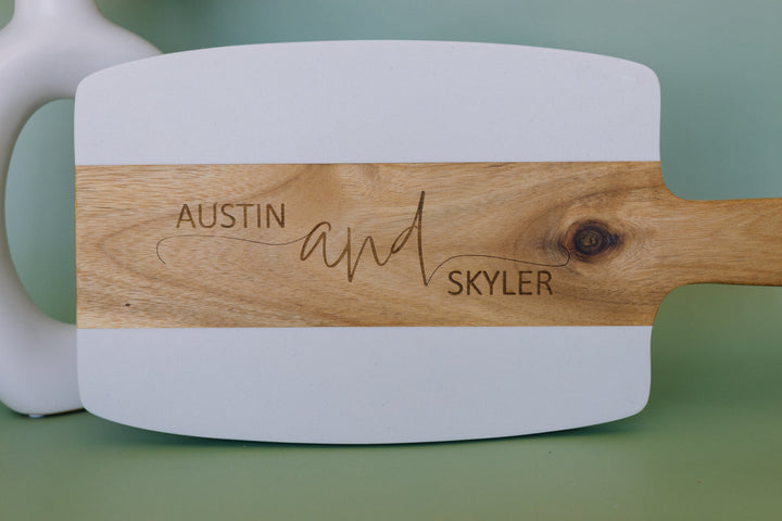 White cutting board with wooden handle engraved with 'Austin and Skyler' on a green background